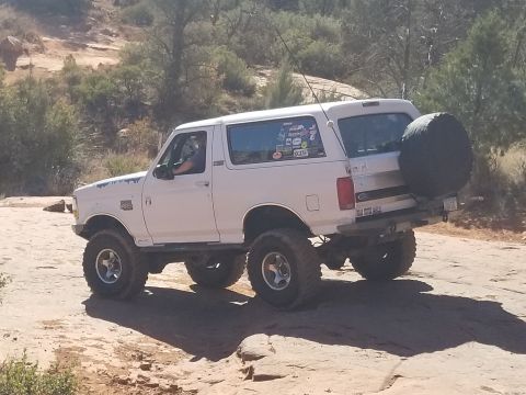 1995 Ford Bronco - A lifted white 1995 Ford Bronco is shown from the rear-three-quarter angle on a dirt road with rocky terrain and sparse vegetation. The veh…