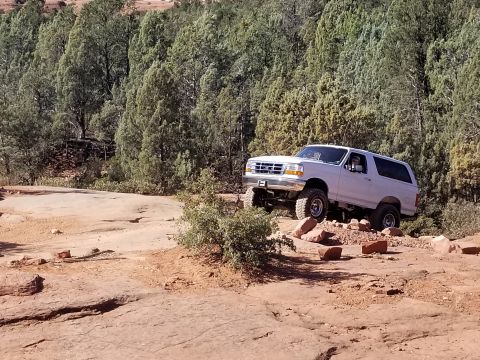1995 Ford Bronco - A white Ford Bronco is driving off-road on a rocky, dirt trail with trees in the background. The vehicle is angled to the right, showing it…