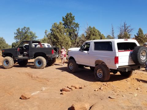 1995 Ford Bronco - Two modified Ford Broncos are parked on a dirt trail with trees and a clear blue sky in the background. The Bronco on the left is black wit…