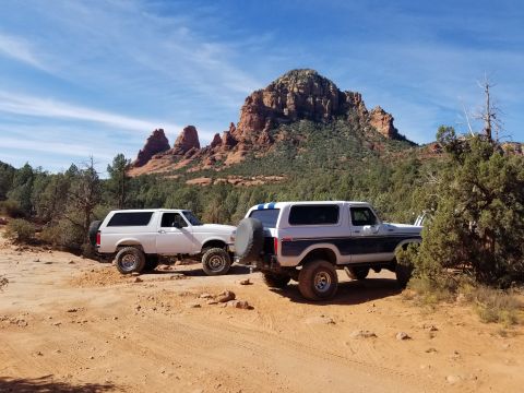 1995 Ford Bronco - Two Ford Broncos are parked on a dirt trail with red rock formations and green trees in the background under a blue sky with scattered clou…