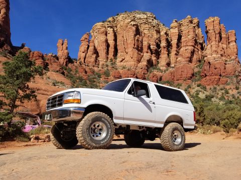 1995 Ford Bronco - A white 1995 Ford Bronco is parked on a dirt road with large red rock formations in the background. The SUV has large off-road tires and a…