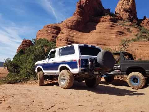 1995 Ford Bronco - A white and blue Ford Bronco with off-road tires is parked on a dirt trail in front of red rock formations. A black pickup truck is parked…