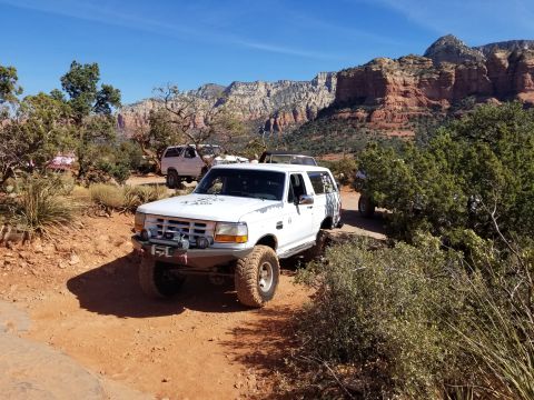 1995 Ford Bronco - A white Ford Bronco is parked on a dirt trail in a desert landscape with red rock formations in the background. The vehicle has large off-r…