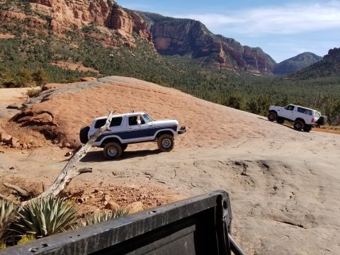 1995 Ford Bronco - Two classic Ford Broncos are off-roading on a rocky, desert landscape with red rock formations and green trees in the background. The Bronc…