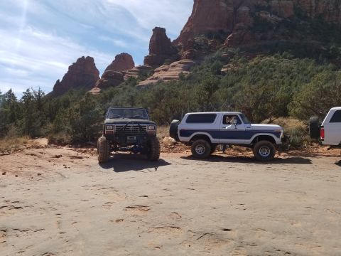 1995 Ford Bronco - Two lifted Ford Broncos are parked on a dirt road with red rock formations and green trees in the background. The Bronco on the left is dar…