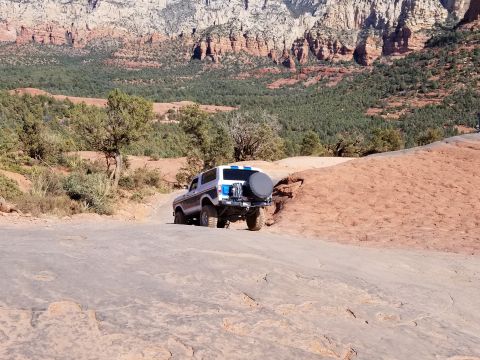 1995 Ford Bronco - A white Ford Bronco with blue accents is driving on a rocky, off-road trail with red rock formations and green trees in the background.