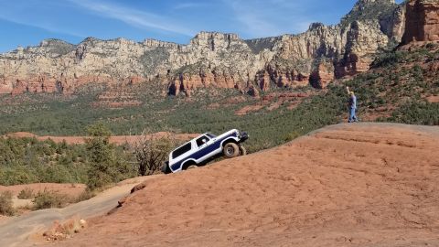 1995 Ford Bronco - A blue and white Ford Bronco is driving up a steep, rocky, red dirt incline with a person standing on a ledge in the background. The vehicl…