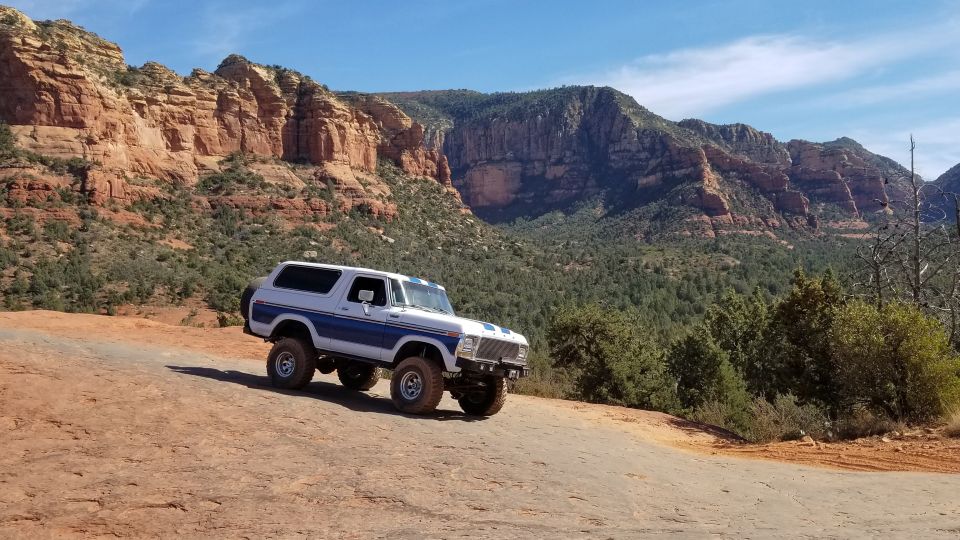 Lifted white and blue Ford Bronco on a dirt trail with red rock canyon background.