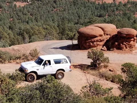 1995 Ford Bronco - A white 1995 Ford Bronco is driving on a dirt road in a desert landscape with red rock formations and green trees in the background.