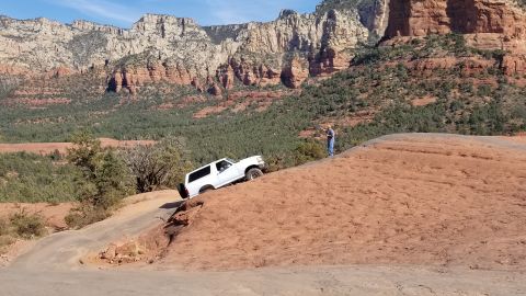 1995 Ford Bronco - A white 1995 Ford Bronco is navigating a steep, rocky incline on a dirt trail with a person standing on the ridge above. The vehicle is pos…