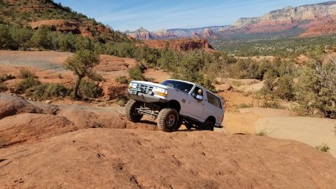 1995 Ford Bronco - A white 1995 Ford Bronco is navigating a rocky, off-road trail with a driver visible behind the wheel. The vehicle is angled on a steep inc…