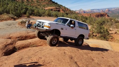 1995 Ford Bronco - A white Ford Bronco is shown off-roading on a rocky, desert terrain with red rock formations in the background. The vehicle has large tires…
