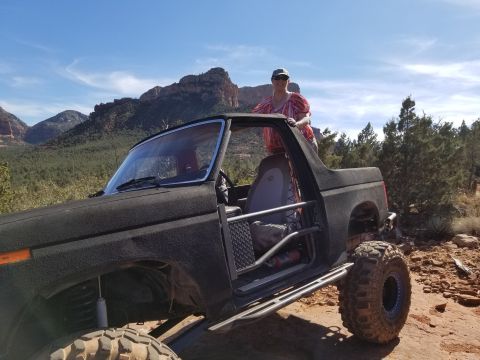 1995 Ford Bronco - A woman stands in the open doorway of a black, lifted Ford Bronco with large off-road tires, parked on a rocky trail with red rock formatio…