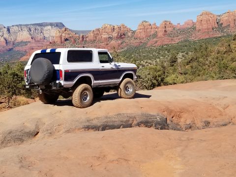 1995 Ford Bronco - A white and dark blue two-door SUV with off-road tires is parked on a rocky, desert trail with red rock formations and green vegetation in…