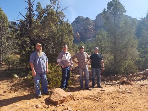 1995 Ford Bronco - Four men stand in a desert landscape with trees and rock formations in the background. The ground is sandy and red, with some rocks and fal…