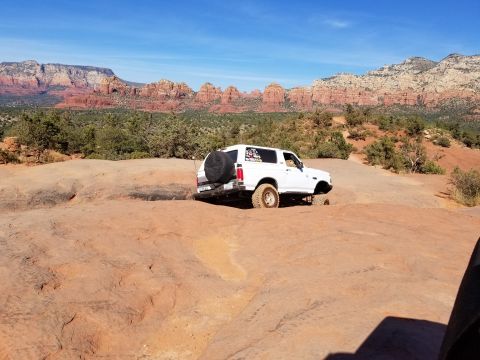 1995 Ford Bronco - A white 1990s Ford Bronco is parked on a rocky, red-rock desert landscape with mountains in the background under a clear blue sky. The vehi…