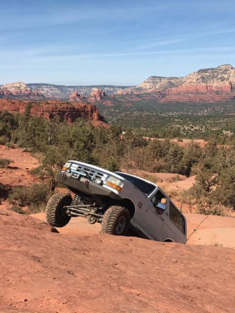 1995 Ford Bronco - A silver 1995 Ford Bronco is shown off-roading on a steep, rocky, red dirt trail with a scenic desert landscape in the background.