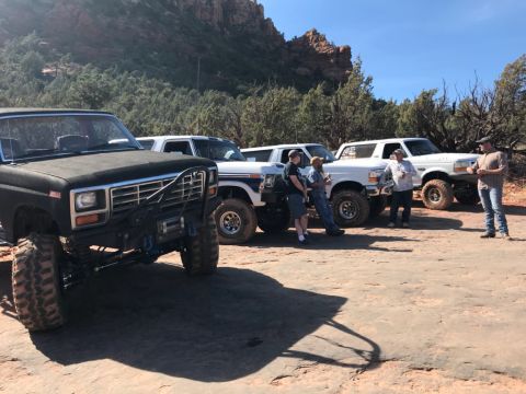 1995 Ford Bronco - A group of people stand around several lifted Ford Broncos and a black Ford pickup truck on a rocky, desert terrain with red rock formation…