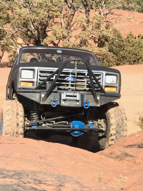 1995 Ford Bronco - A modified black Ford Bronco is shown from the front, parked on a rocky, desert terrain with sparse vegetation in the background. The vehic…