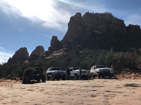1995 Ford Bronco - Four off-road vehicles are parked on a rocky, desert terrain with large red rock formations and trees in the background under a partly clou…