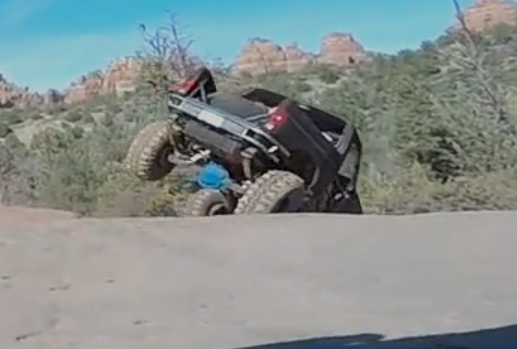 Heavily modified black off-road truck tilted on a dirt hill with front tires in the air.