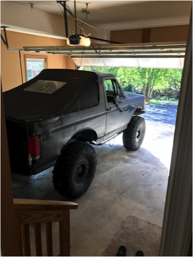 1989 Ford Bronco - A lifted, black Ford Bronco with a soft top is parked inside a garage. The vehicle features large, off-road tires and is viewed from the re…