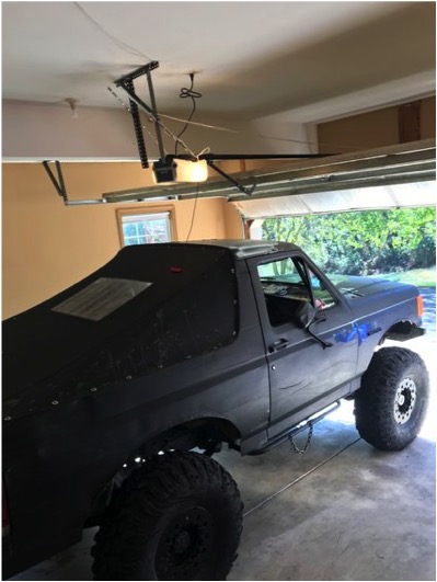 1989 Ford Bronco - A lifted 1989 Ford Bronco with large off-road tires is parked inside a garage with the garage door open. The vehicle has a black soft top a…
