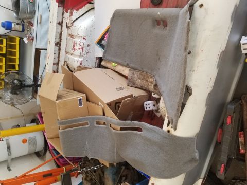 1967 Ford Bronco - A pile of automotive interior trim pieces, including two grey carpeted panels, are stacked with cardboard boxes in a cluttered garage or wo…