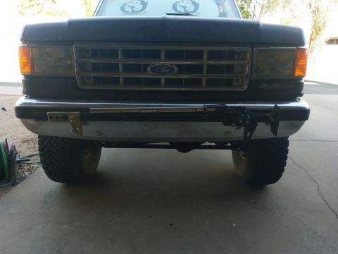 1988 Ford Bronco - Front view of a dark colored Ford Bronco with a chrome bumper mounted with a Hi-Lift jack and a tow hook. The vehicle is parked on a concre…