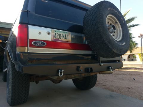 1988 Ford Bronco - The rear view of a dark blue Ford Bronco with a spare tire mounted on the back. The vehicle has a custom black bumper with tow hooks and a…