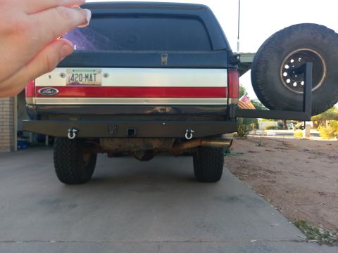 1988 Ford Bronco - The rear of a dark colored SUV with a spare tire mounted on a swing-away carrier. The vehicle has a custom bumper with tow hooks and a lice…