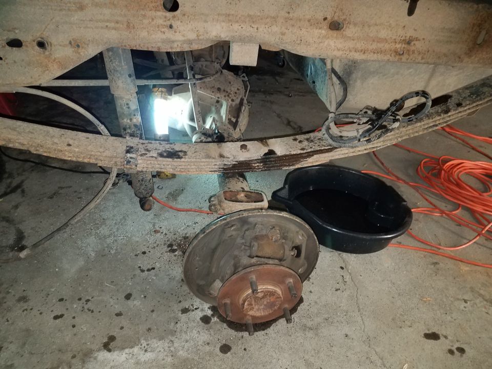 Underside of a vehicle showing a leaf spring, rear axle, and brake drum assembly. A drain pan with fluid and orange extension cords are nearby