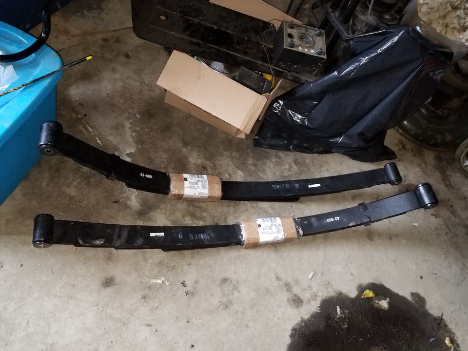 Two black leaf springs with part numbers on a concrete floor.