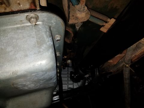 1996 Ford Bronco - The underside of a vehicle shows a silver-colored oil pan with several bolts securing it. Further back, a black metal component, possibly a…
