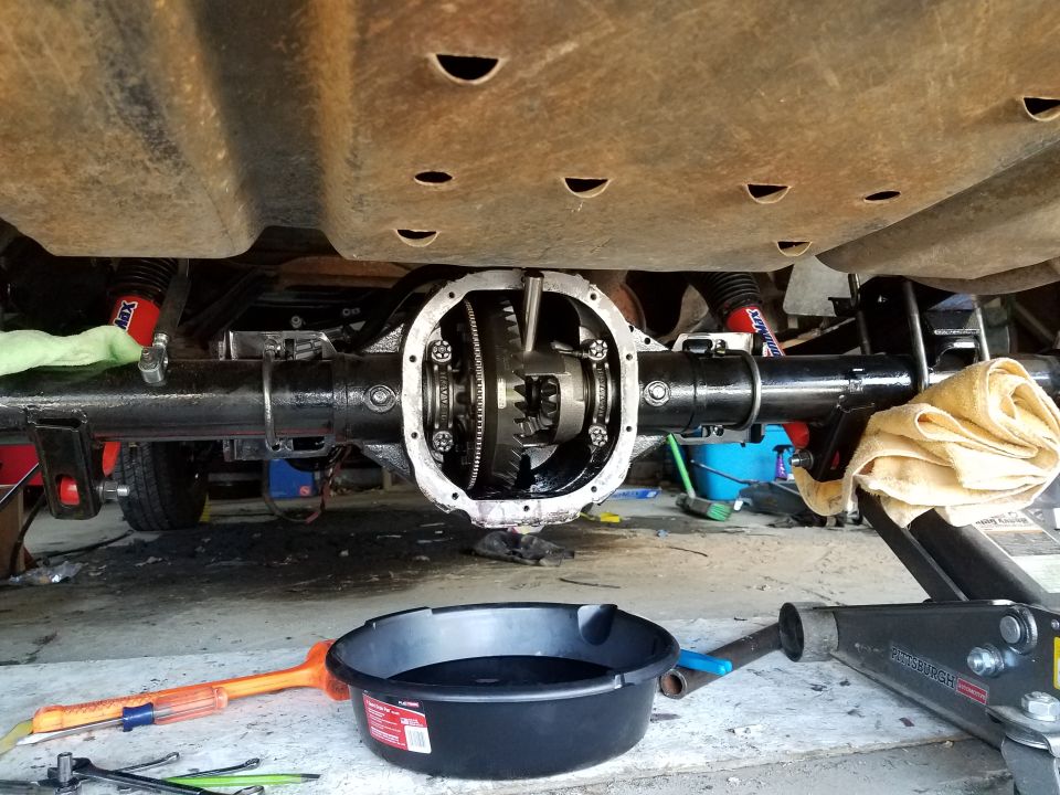 Rear differential of a vehicle with cover removed, showing gears. Tools and drain pan below.