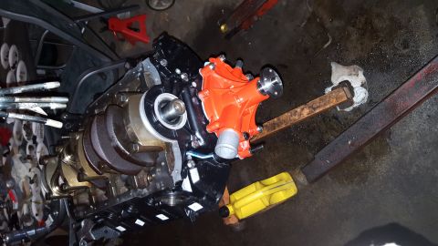 1996 Ford Bronco - An engine block with a bright orange water pump installed is shown on a garage floor. The crankshaft is visible on the left side of the eng…