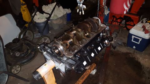 1996 Ford Bronco - A black V8 engine block with a crankshaft installed is on an orange engine stand. Various tools and equipment, including a lawnmower and a…