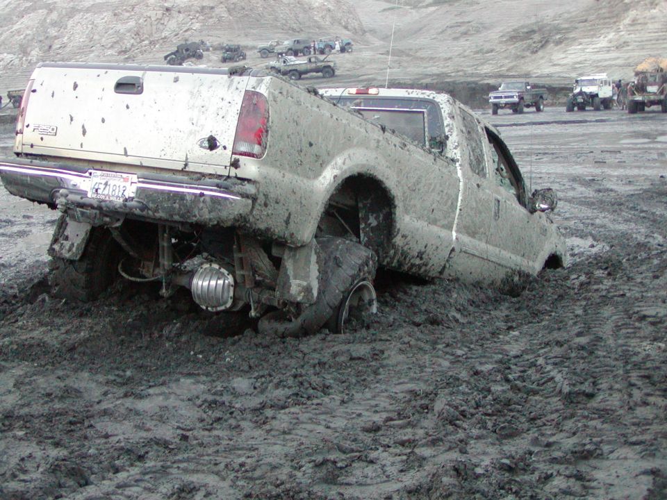 White Ford Super Duty pickup buried nose-down in deep mud, rear axle and tire exposed, at an off-road area.