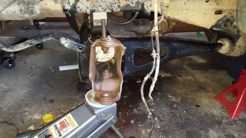 1996 Ford Bronco - A vehicle's suspension components are shown in a garage setting. A floor jack supports a rusty bracket, with brake lines visible nearby.