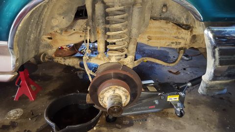 1996 Ford Bronco - The front suspension and brake rotor of a vehicle are visible, supported by a red jack stand and a hydraulic floor jack. A black oil drain…