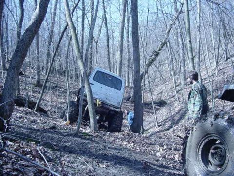 1992 Ford Bronco - A white SUV is stuck in the mud on a wooded trail, with a person standing nearby observing the situation.