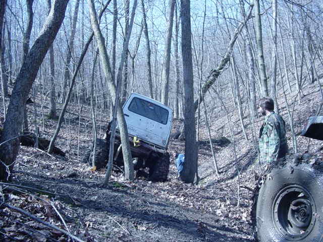 White SUV stuck in mud on wooded trail, driver standing nearby