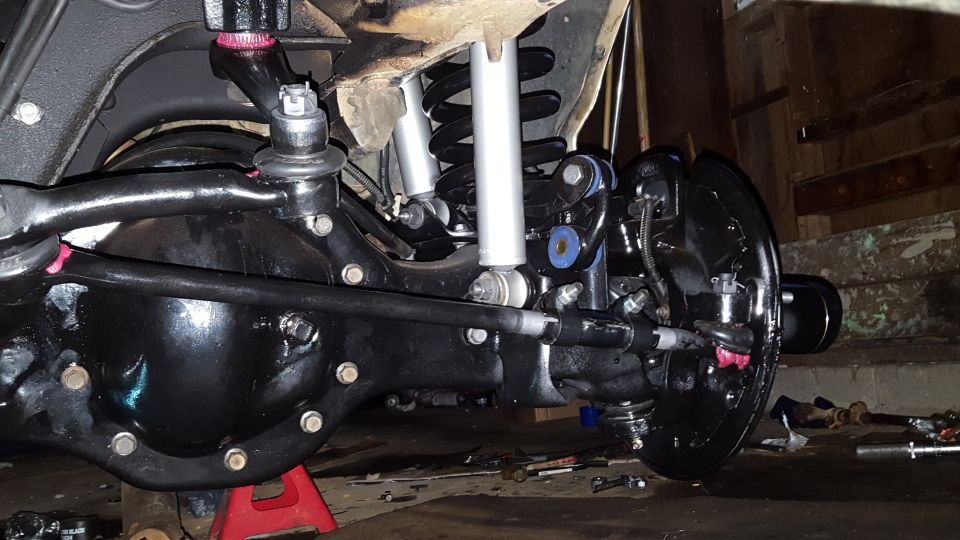 Underside of vehicle suspension and drivetrain with black differential, silver shocks, and red jack stand.