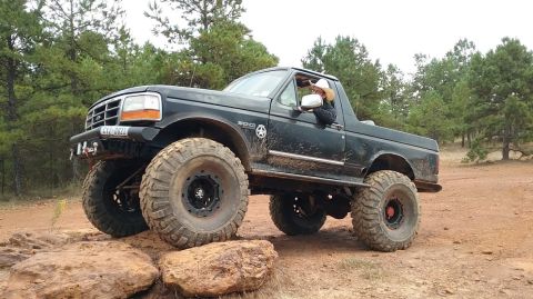 1995 Ford Bronco - A black, lifted 1995 Ford Bronco with oversized off-road tires is shown navigating a rocky trail in a wooded area. The driver, wearing a co…