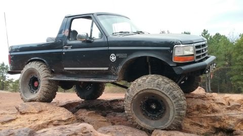 1995 Ford Bronco - A black 1995 Ford Bronco is shown off-road, navigating large rocks with its oversized tires. The vehicle has been modified for off-road use…