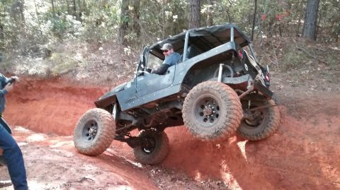 1995 Ford Bronco - A dark-colored Jeep Wrangler is navigating a steep, red dirt incline with its front wheels lifted high off the ground. The driver, wearing…