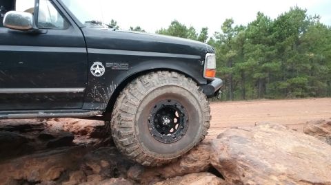 1995 Ford Bronco - The front passenger side of a black Ford Bronco is shown angled over large rocks on a dirt trail. The vehicle has aggressive off-road tires…