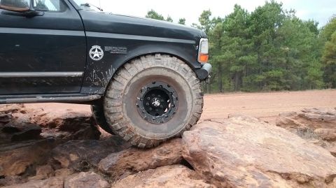 1995 Ford Bronco - The front driver's side of a black Ford Bronco is shown angled over large rocks, with its large, off-road tire prominently featured. The ve…
