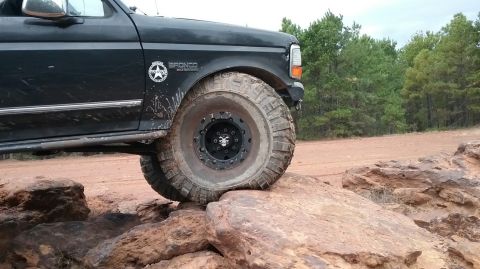 1995 Ford Bronco - The front passenger side of a black 1995 Ford Bronco is shown driving over large rocks on a dirt road. The vehicle has large off-road tires…