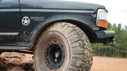 1995 Ford Bronco - A close-up, low-angle shot of the front fender and large, aggressive off-road tire of a black Ford Bronco. The vehicle is on a dirt road wi…